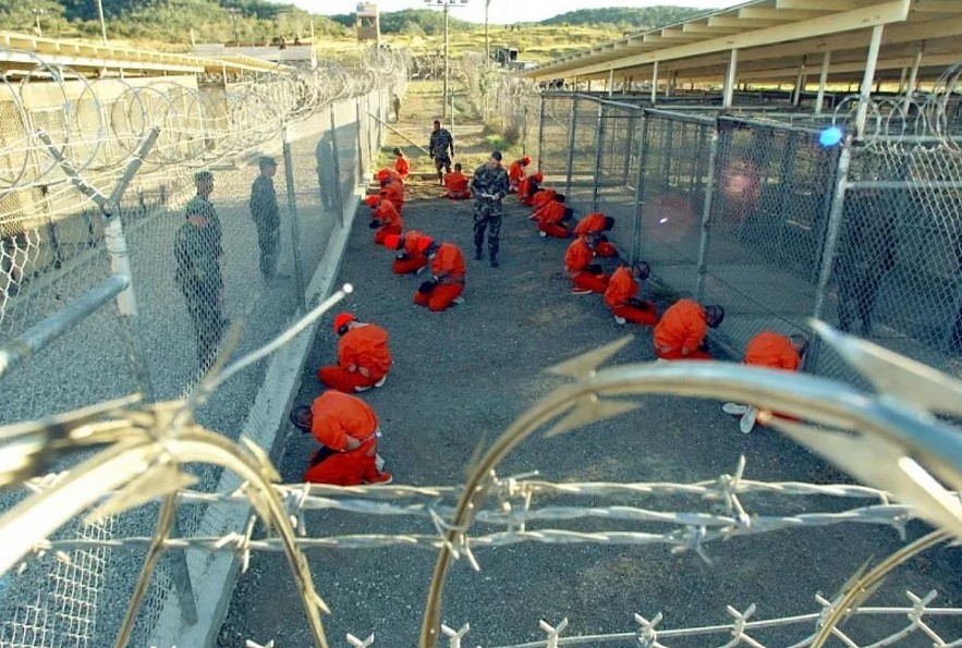 In this official handout photo from the U.S. Navy, U.S. Military Police oversee Taliban and al-Qaeda detainees dressed in orange jumpsuits on January 11, 2002, as they undergo in-processing at Camp X-Ray, a temporary detention facility at Naval Base Guantanamo Bay, Cuba.